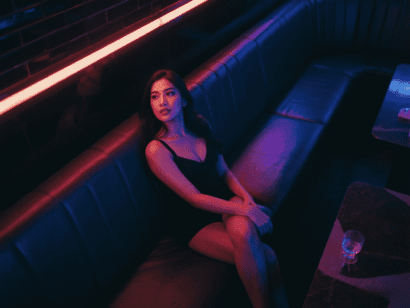 Woman in black dress on a booth in a dimly lit club with neon lights.